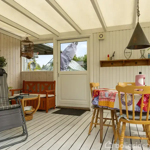 En hyggelig veranda indeholder et bord med farverig dug, omgivet af stole. Til venstre står en liggestol ved siden af en plante. Væggen har en hylde med dekorationer.