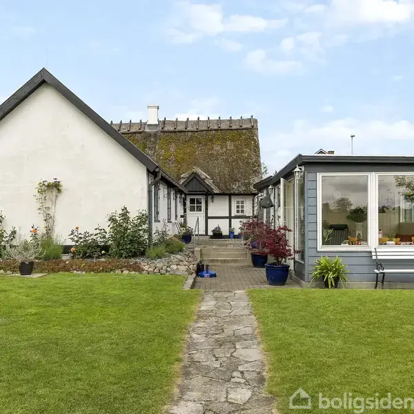 Huset med stråtag og hvid facade står stille i en velplejet have med grøn græsplæne og blomsterkrukker. Stensti fører op til indgangen under en blå himmel.