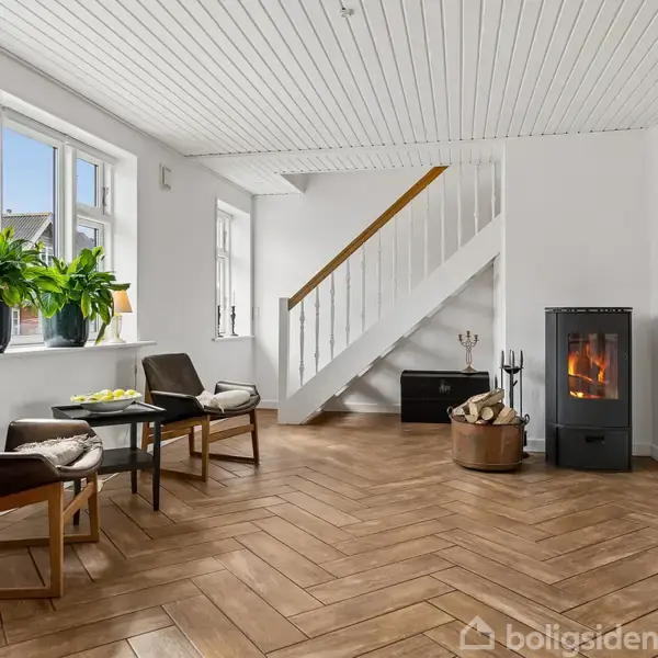 To lænestole står omkring et lille bord i et lyst rum med trægulv. En brændeovn er i hjørnet ved siden af en trappe. Store vinduer med planter som udsigt.