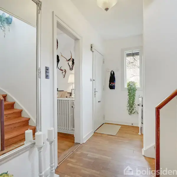 Et lyst gangområde med en stor spejl i dekoreret ramme. Trætrapper fører opad. Grønt vedhængende plante ses tæt på vinduet, og væggen har enkelte kroge.