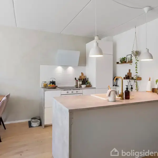 Et lyst køkken med køkkenø har en vask og planter på bordpladen. To lamper hænger over øen. I baggrunden står et spisebord med blomster.