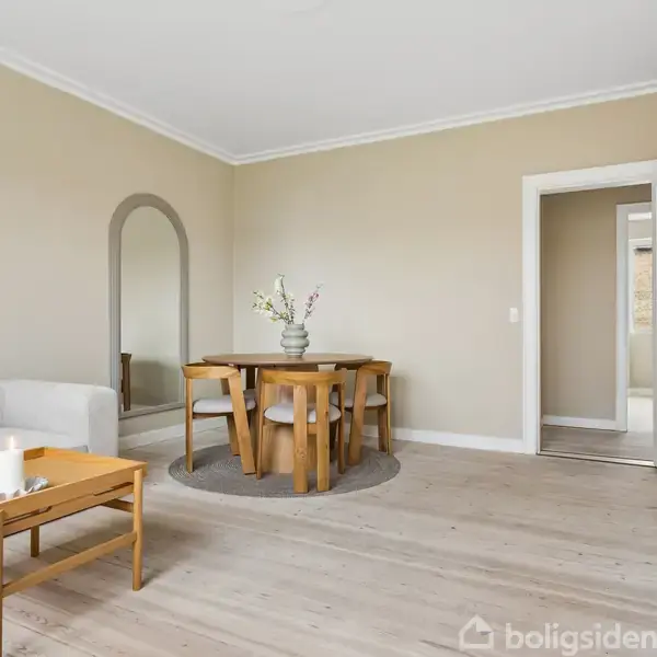 Sofa og sofabord står i en stue med lyse trægulve. Et rundt spisebord med stole er mod væggen. Lys beige vægmaling komplementerer det enkle, moderne design.