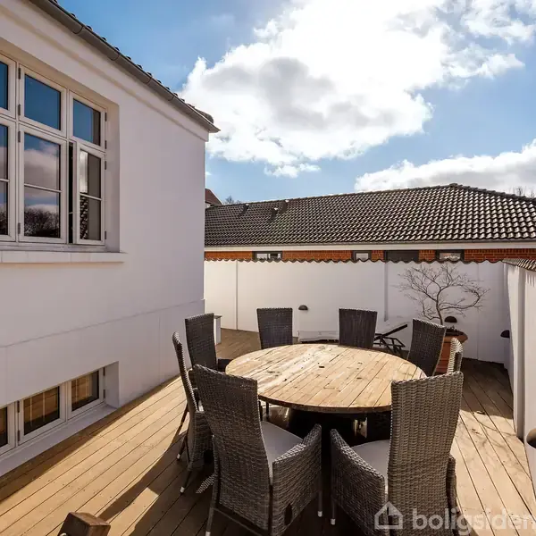 Rundt havebord omgivet af otte kurvestole står på en træterrasse ved siden af et hvidt hus med store vinduer. I baggrunden ses en hvid mur og et tag med røde tegl.