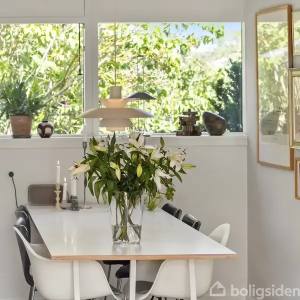 Badeværelse med hvidt skab under en stor spejl, omgivet af rammer med portrætter. Lysindfald fra vindue med planter. Gråt gulv og bruseområde bag beige forhæng.