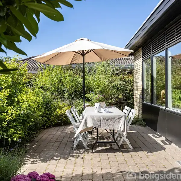 En udendørs terrasse med et bord og stole står under en parasol. Bordet er dækket med en dug, og omgivet af grønne planter og et hus i baggrunden.