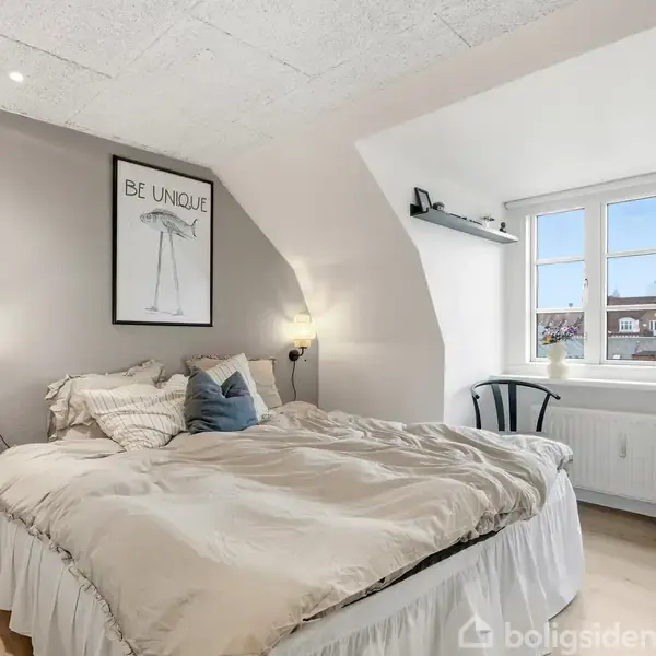 Seng med puder står i et lyst soveværelse. Plakat over sengen viser teksten "BE UNIQUE". Der er en stol ved vinduet med udsigt til hustage.