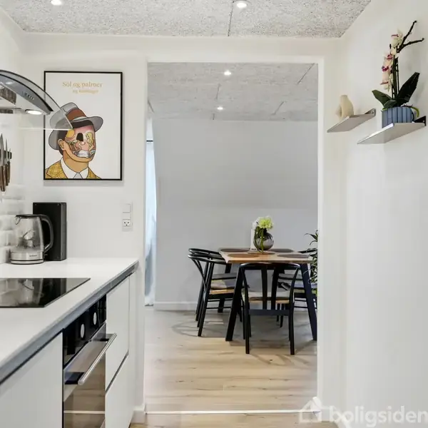 Et moderne køkken med hvide overflader og indbygget kogeplade. Til venstre hænger et billede af en person med teksten "Lad os så komme i gang". I baggrunden ses et spisebord med blomster.