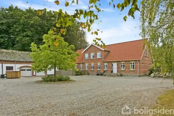 En stor, rød murstensbygning med rødt tegltag står i en grusbelagt gårdsplads. Til venstre ses en garage, omgivet af grønne træer i baggrunden.