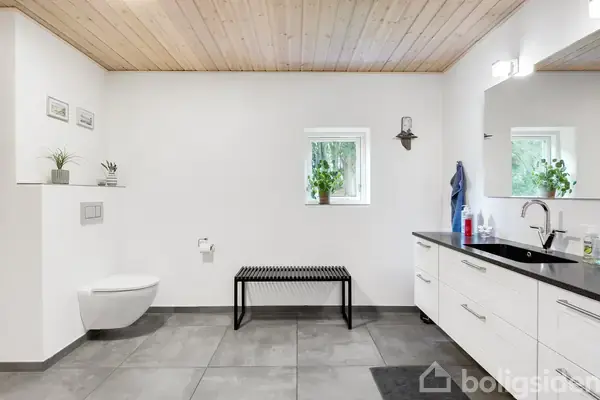 Toilet er monteret på en hvid væg i et moderne badeværelse. Sorte fliser dækker gulvet, en bænk står ved siden af, og et lille vindue med planter tilfører lys.