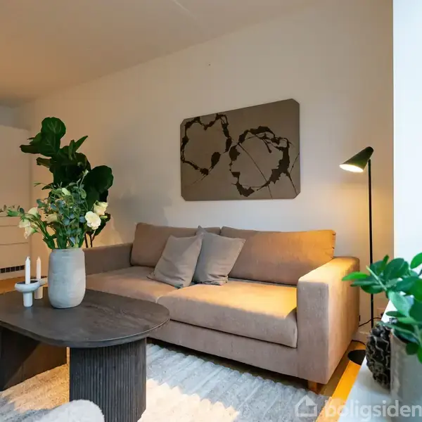 Brun sofa står i en moderne stue med et vægmaleri bagved. Foran sofaen er et mørkt sofabord med en vase med blomster. Køkkenet ses i baggrunden til venstre.