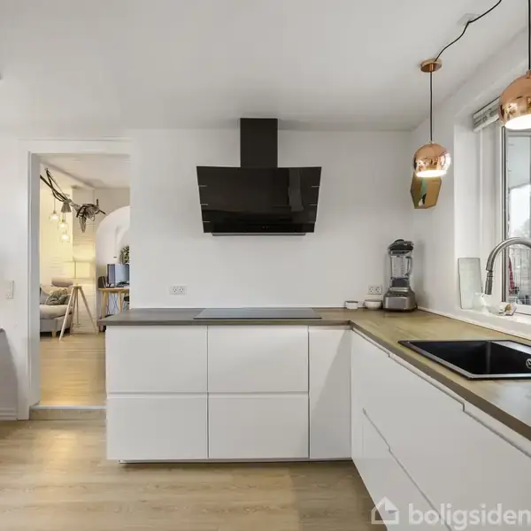Køkken med hvide skabe har en sort emhætte og blender på bordpladen. To kobberfarvede lamper hænger over vasken. Spiseplads i baggrund. Lyst rum med trægulv.