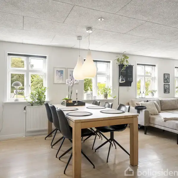 En spisestue med et langt træbord og sorte stole står ved siden af en hjørnesofa. Vinduer i baggrunden giver lys og udsigt til haven, med planter på vindueskarmen.