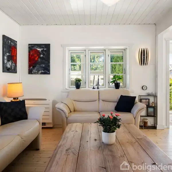 Stue med lyse lædersofaer omkring et træbord; to malerier hænger på væggen. En lampe og planter pynter rummet, hvor store vinduer giver udsigt til en grøn have.