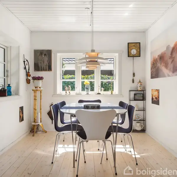 Spisebord med fire stole står i en lys spisestue. Lampen hænger over bordet. Væggene er dekoreret med billeder og hylder, og vinduer lukker dagslys ind.