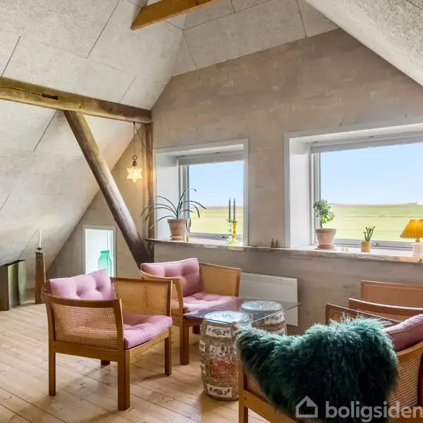 Stue med skrå loft og synlige bjælker. Fire polstrede stole står omkring et lavt bord. Vinduer viser landskab udenfor. Grøn plante, orange lampe og vaser står i vindueskarmen.