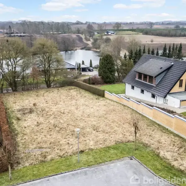 Grunden er ubebygget med vild bevoksning, beliggende ved siden af et moderne to-etagers hus med sort tag. I baggrunden ses en sø omgivet af træer og åbne marker.