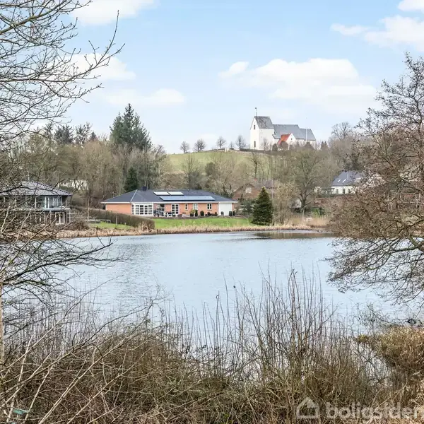 En sø omgivet af træer og buske ligger i forgrunden, mens huse og en kirke med hvid facade står på en bakke i baggrunden under en blå himmel med skyer.