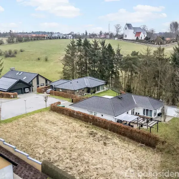 Huse med mørke tage står i en boligområde omgivet af træer og marker. I baggrunden ses en kirke på en bakke under en blå himmel med skyer.