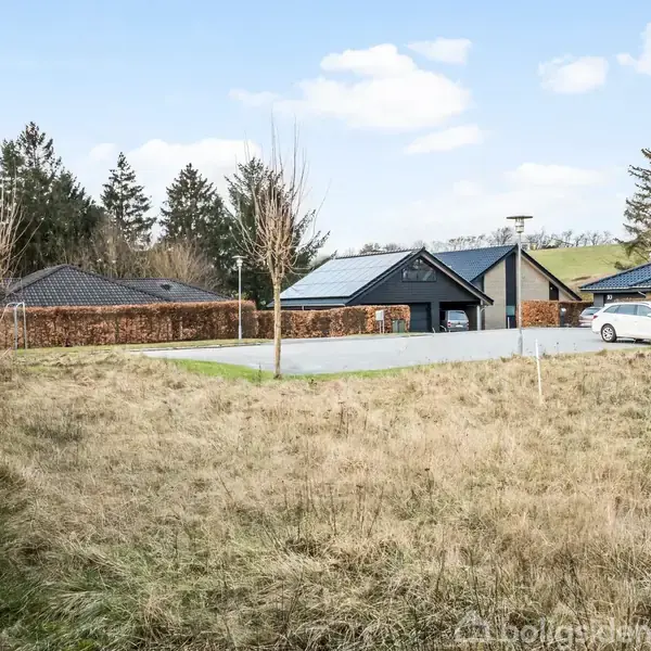 Græsplæne strækker sig forbi en p-plads med enkelte unge træer. Bagved ses sorte hustage, en hvid bil og træer under en blå himmel med få skyer.