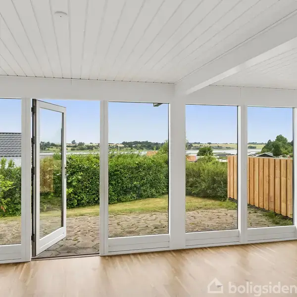 Glasdør åbner ud til en stenbelagt terrasse omkranset af frodig bevoksning og udsigt over et landligt landskab. Indendørs er lyst trægulv og hvidt loft.
