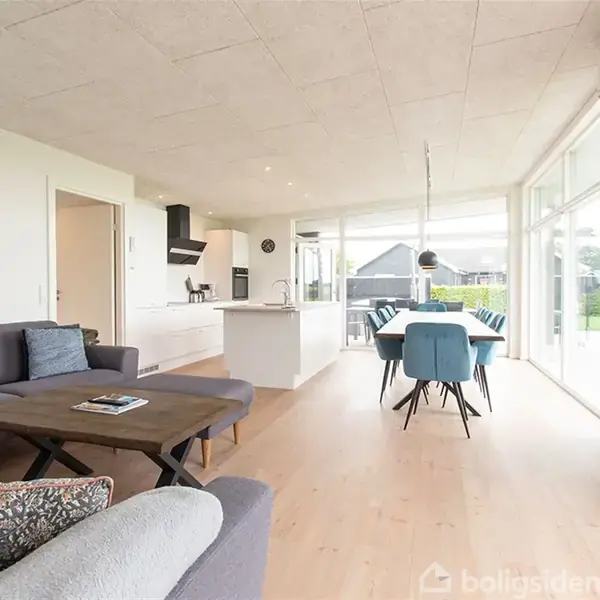 Sofaen står i en lys stue med åbent køkken, hvor et trægulv fører til store vinduer. Et spisebord med blå stole er i midten, og en vægmonteret tv-skærm ses.