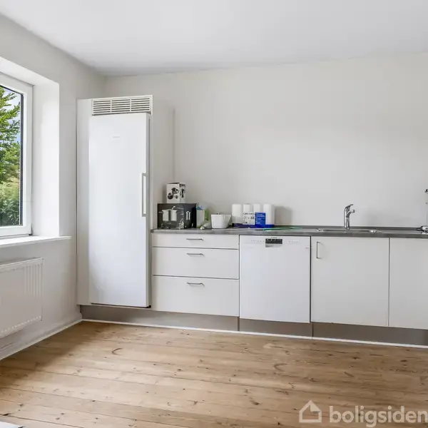 Lyse hvide køkkenelementer med håndtag fylder en væg under et vindue. En kaffemaskine og brødrister står på bordet. Rummet har et trælaminatgulv og udsigt til en grøn have.