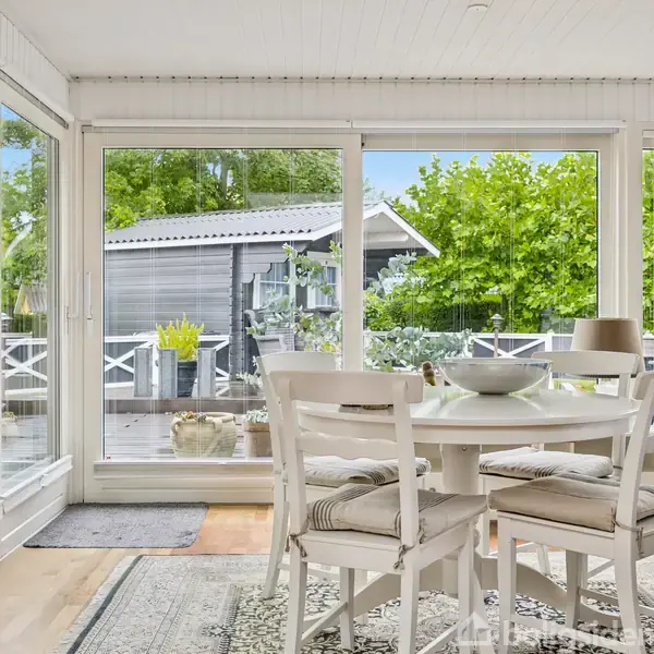 Et rundt spisebord med fire stole står i en vinterhave, omgivet af store vinduer. Udenfor ses en terrasse med planter og et lille skur, omkranset af grønne træer.