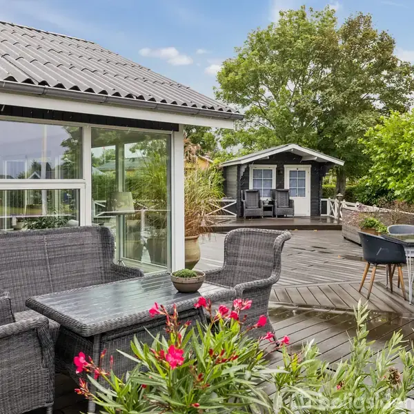 Terrassemøbler står på en træterrasse ved siden af et hus med store vinduer. En parasol er opstillet ved et rundt bord, omgivet af buske og et lille haveskur i baggrunden.