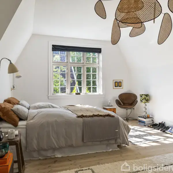 Soveværelse med dobbeltseng placeret under et skråloft. Seng med sengebord ved siden af. Stor vindue bagved slipper naturligt lys ind. Hylder med sko ved radiator. Snegleformet loftslampe.