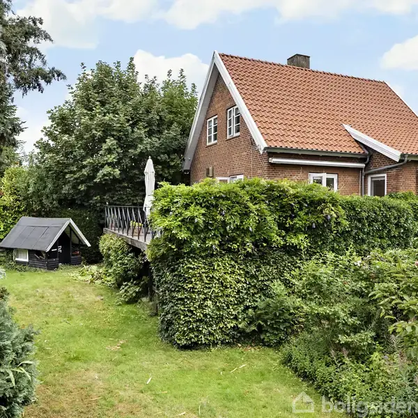 Et rødt murstenshus med rødt tegltag, omgivet af en frodig have med buske og træer. En terrasse med havemøbler og en parasol er placeret ved huset.