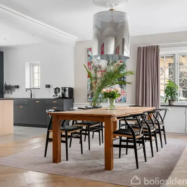 Et træspisebord med fire sorte stole omkring det står i en lys spisestue. En vase med blomster pryder bordet. Køkkenområdets grå skabsenheder ses i baggrunden ved et vindue.