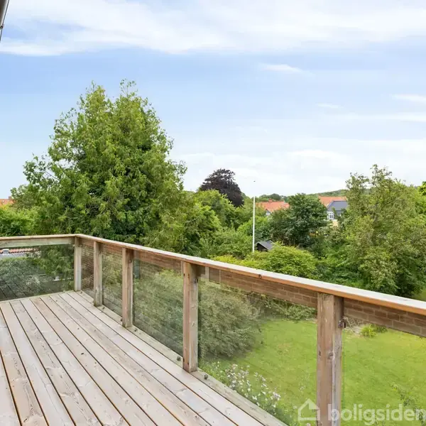 En træterrasse strækker sig langs et murstenshus, omgivet af en frodig have med grønne træer. Udsigten viser en rolig, naturskøn baggrund under en klar himmel.