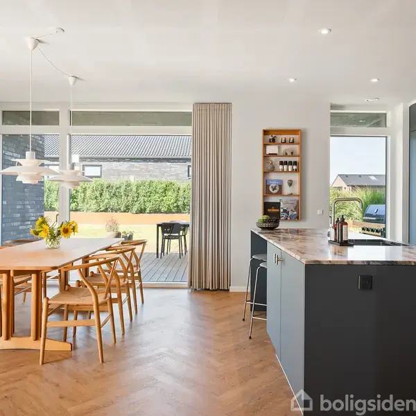 Spisebord med træstole står i et lyst køkkenalrum. Foran glasskydedøre, er en terrasse synlig udenfor. Billedet inkluderer moderne lamper, køkkenø og indbyggede skabe.