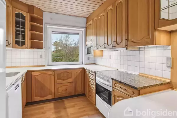 Et køkken med træskabe og hvide fliser. Der er en ovn og komfur, et stort vindue med udsigt til naturen udenfor. Trægulve og loft i samme stil.