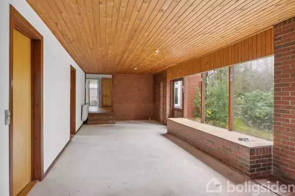 En tom korridor med trægulv- og loft, en murstensvæg med store vinduer, der vender ud mod en have. Åbne døre fører til andre rum, og naturligt lys fylder rummet.