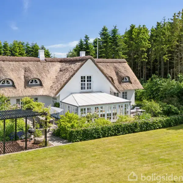Et stråtækt hus står omgivet af frodig have og skov. En pergola er placeret ved siden af huset, som har en stor græsplæne foran.