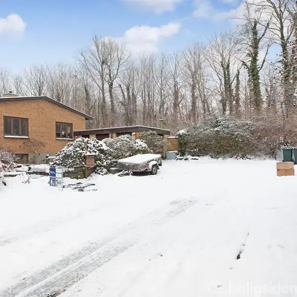 Et hus i mursten står dækket af sne i en vinterlig have omkranset af træer. I forgrunden ses en snedækket indkørsel og nogle affaldsbeholdere. Der er et til salg-skilt foran huset.