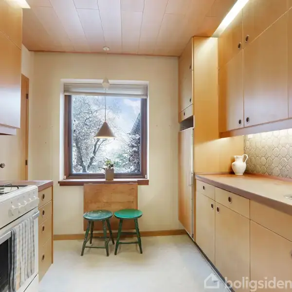 Et køkken indeholder brune skabe og en hvid komfur. To grønne taburetter står ved et lille bord under et vindue med sne udenfor. En hvid kande står på bordet.