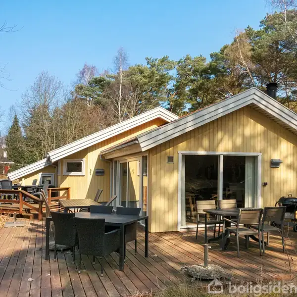 Et gulmalet sommerhus med store vinduer står på en træterrasse. Møbler, herunder stole og et bord, er placeret udenfor. Omgivet af træer og skovområde.