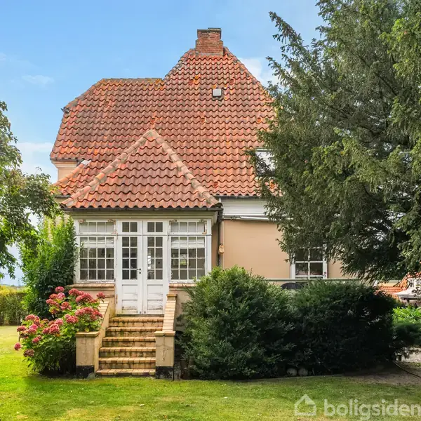 Et hus med rødt tegltag står midt i en have. Trappe fører op til en hvid hoveddør. Omgivet af buske og træer under en blå himmel.