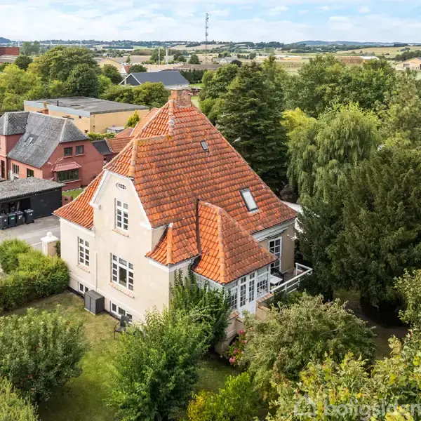 Et stort, hvidt hus med rødt tegltag står omgivet af træer og tæt på andre bygninger i et grønt landskabsområde.
