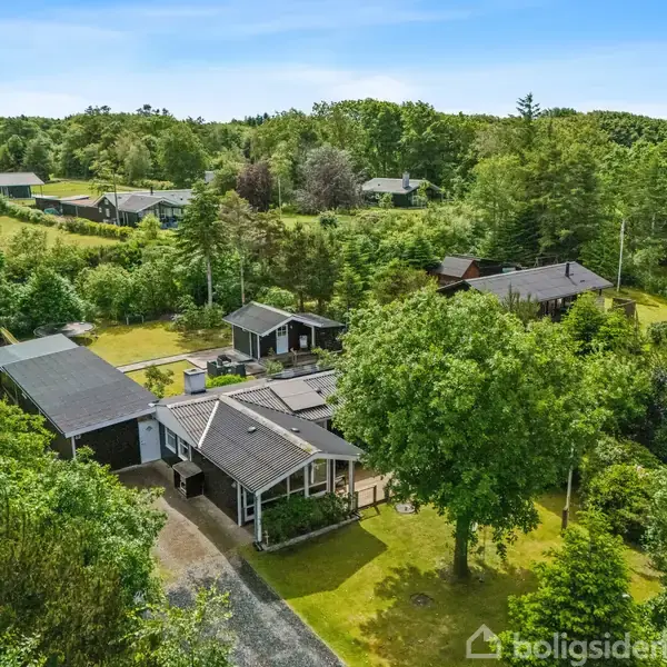 Et hus omgivet af frodig vegetation ligger i et grønt område med flere spredte huse. Haven er velplejet, og der er en opkørsel fører til garagen.