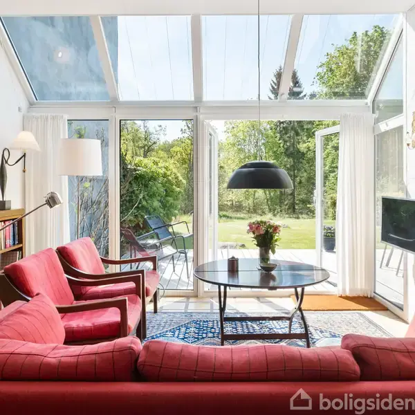 Stue med røde sofaer og et rundt bord. Store vinduer åbner ud til en grøn have. Hylder med bøger og lamper skaber en hyggelig atmosfære.