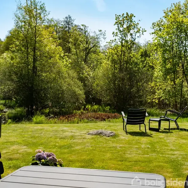 Havebord står på en græsplæne med to stole ved siden af, omgivet af frodig have. I baggrunden ses tætte træer og buske under en blå himmel.