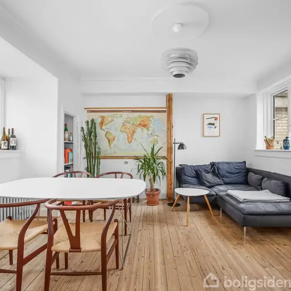 Et spisebord er omgivet af stole i en lys stue med trægulv. Til højre er en mørkegrå hjørnesofa. En vægdekoration forestiller et verdenskort, omgivet af planter.