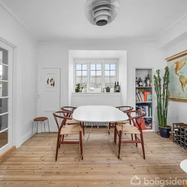 Et spisebord med seks stole placeret i et lyst rum med trægulv. En stor verdenskort hænger på væggen, omgivet af planter og en bogreol. Glasdøre fører til et tilstødende rum.