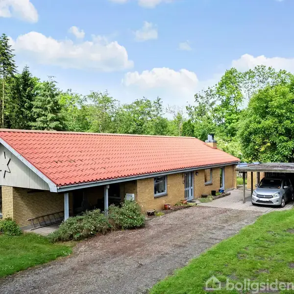 En gul murstensvilla med rødt tegltag står omgivet af frodige træer, en græsplæne og en grussti foran huset. En bil er parkeret under en carport til højre.