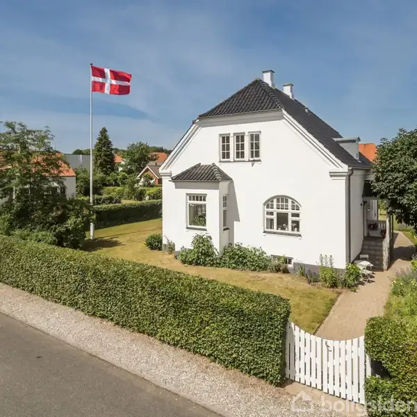Hvidt hus med mørkt tag står i en have med hæk og græsplæne. En dansk flagstang ved siden, omgivet af træer og andre huse på en stille vej.