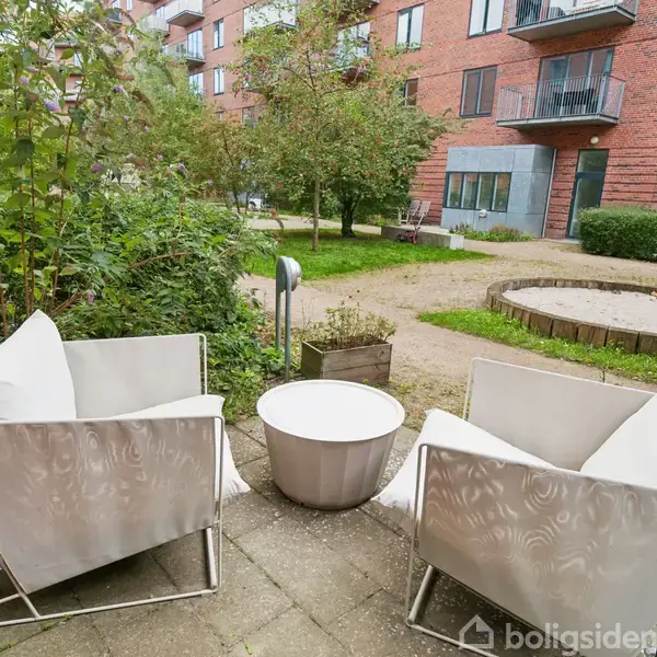 Køkken med hvide skabe står langs en væg. Kaffemaskiner og indbygget ovn er placeret på en bordplade. Rummet har lyse træparketgulve og hvide vægge.