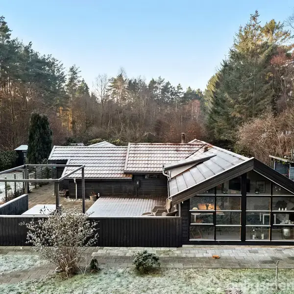 En sort træhytte står stille i en skovrig baggrund med tætbeplantede træer. Terrassen foran huset er omkranset af et lavt træhegn, og himlen er klar over grunden.